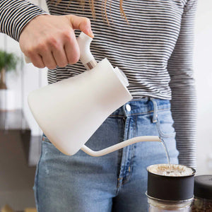 Hånd, der hælder vand fra en hvid elkedel med smal tud ned i et kaffefilter, del af EspressoGear, alt om kaffe.
